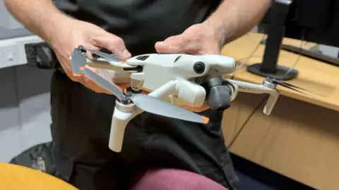 A light grey gimballed camera drone with four arms, each with two propellors. It is being held in a police officer's hands.