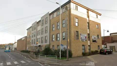 A four-storey accommodation block on a street in day time. There is a bus in the distance approaching a zebra crossing and a parked car to the right of the building.