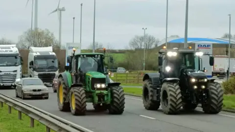 Two tractors drive down a dual carriageway as traffic starts to build up behind them. 