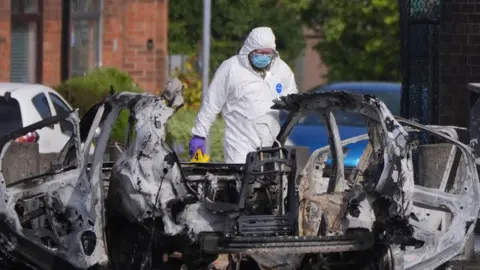 Forensic officer, in white suit, and blue face covering, beside the debris of a car after an explosion in Dunmurry. 