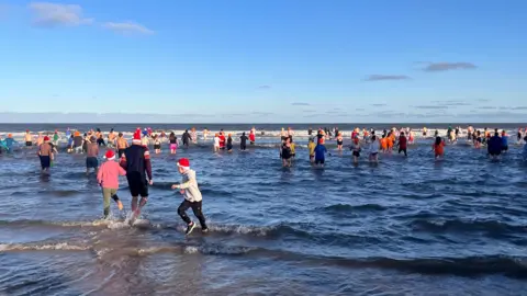 Proyecto Faro. Una multitud en el mar con un cielo azul casi despejado. Numerosas personas llevan gorros de Papá Noel y colores brillantes.