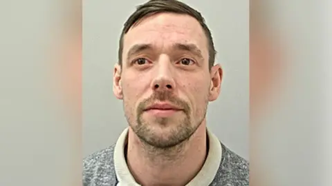 Police custody image of Shaun Fuller who has brown eyes and brow hair with brown stubble. He is wearing a cream coloured grey jumper. 