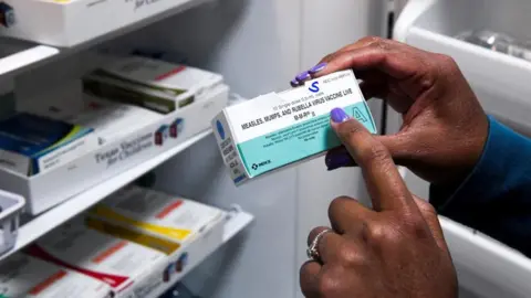 A box containing the vaccine against measles, kanthamala and rubella is being conducted by a woman with her right hand, pointing to writing on it with the index finger on the left side.