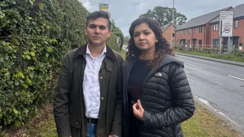 A couple, perhaps in their 40s, stand on the side of a road in a semi-rural area on a cloudy day. They are wearing jackets and are standing on a grass verge, bordered by a hedge. There is a bus stop sign behind them. On the other side of the road is a line of new-build houses and a road sign. The road is wet.