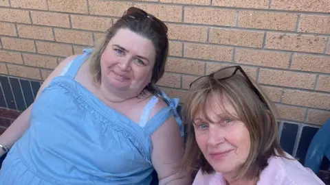 Rona McClure and Margaret Clark sitting outside, against wall. They are smiling into the camera.