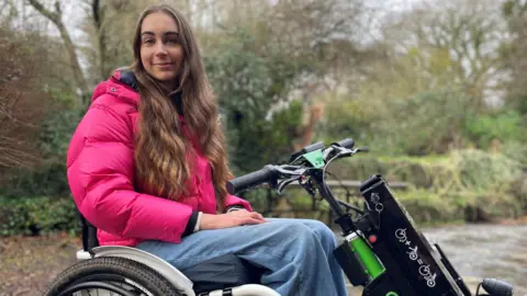 BBC Bethany Handley sitting in her wheelchair with a power attachment looks at the camera while outdoors