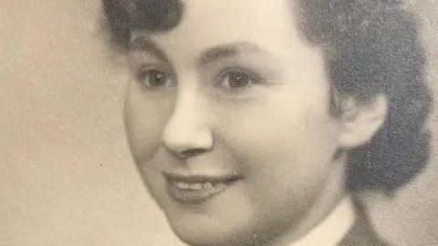 Jan Hall A young woman in uniform smiles in a sepia toned photograph. She has curly hair which looks dark, falling just above her collar, and a fringe.