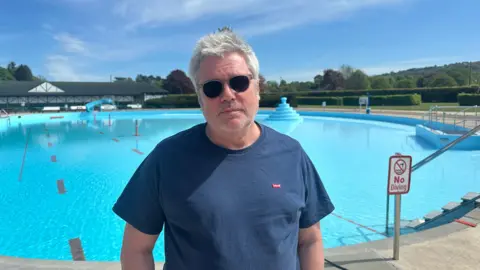 Jessica Bayley/BBC A man with grey hair wearing Ray-Ban-style sunglasses stands in front of a lido on a sunny day. He is wearing a navy T shirt.
