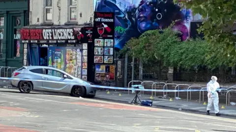 Silver car pushed nose-first against the curb on a road. It is behind a blue and white police tape cordon. Someone in white forensic clothing can be seen in the foreground. An off-licence can be seen in a background, with a dystopian blue-hued mural of a woman on the side.