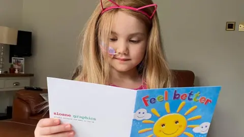Family Emilia is reading a greetings card that says "feel better" on the front. She is wearing her long blonde wig from The Little Princess Trust and has an Alice band with cat ears on it. She has a tube to her nose which is taped to her face as she undergoes treatment.