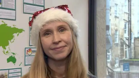 A woman stands in front of a window and a classroom display with countries on and signs. She has blonde hair and is wearing a Christmas hat with red sequins and a fluffy band.