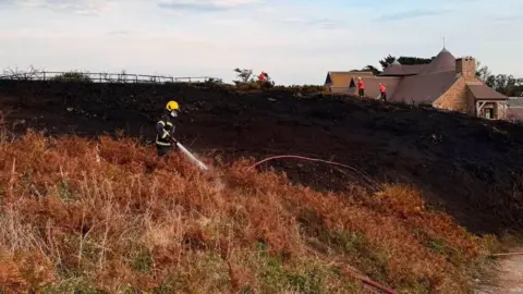 A firefighter sprays water from a hose onto a patch of land which has been burnt during a fire in Jersey. Three other firefighters are doing similar work in the background.
