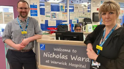 John Blair is wearing a black and white check shirt and black trousers and has a blue NHS lanyard around his neck. He has brown hair and a short beard and is wearing glasses. He is standing to the left of a sign which says welcome to Nicholas Ward at Warwick Hospital. On the other side of the sign is Rachel williams. She is wearing a black dress and a blue NHS lanyard and has mousy blonde hair.