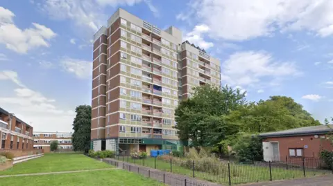 A screenshot from Google street view showing the existing residential tower block. It is a tall brick building with balconies on each floor and a fenced green space at the bottom.