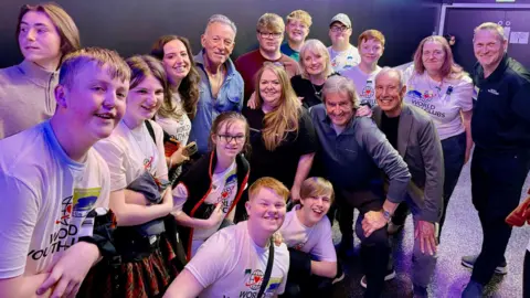 World Youth Clubs Bruce Springsteen stands in the middle of a group of around a dozen teenagers and also adults in a backstage area.