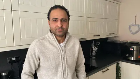 A man in a cream jumper stands in his kitchen with a serious expression