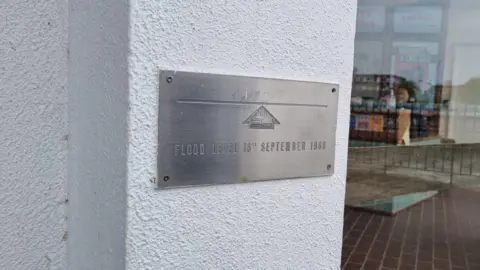 Harrison Galliven / BBC A plaque on a white wall reading: "Flood level 18th September 1968", with an arrow pointing to a line.