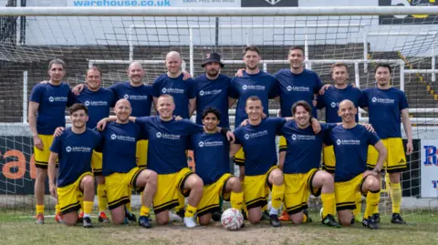 Jon Jerrome A men's football team dressed in kit of blue T-shirt and yellow shorts are pictured kneeling in front of the goal