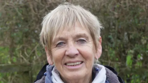 Jamie Niblock/BBC A woman with a blonde pixie hair cut smiles at the camera. Behind her is a hedge. She wears hooped earings with a blue ball on the end of each. She is also wearing a white and purple scarf around her neck. 