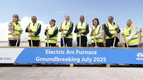 Sam Robinson/BBC Politicians and officials from Tata stand with shovels around a wooden box with "Electric Arc Furnace Groundbreaking July 2025" written across it.
