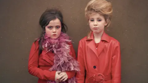 Nia Day as a younger child and Poppy. They are two white girls both wearing all red clothes and coats. Nia, left, has a reddish feather boa around her neck. Both girls have beehive type hairstyles and are made up.
