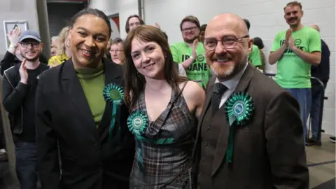 Getty Images Three smiling candidates with Green party rosettes pose in front of clapping activists 