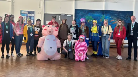 E-ACT Venturers’ Academy A group of teachers stand in a school gymnasium facing the camera. They are all wearing different fancy dress outfits for World Book Day