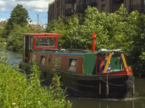 Eileen Martin A red and orange canal boat in the water. Trees and bushes line the canal on both sides.
