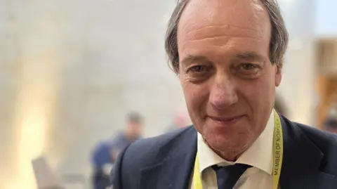 Paul Moseley/BBC Andrew Jamieson is wearing a navy jacket on top of a white shirt and navy tie. He is also wearing a yellow lanyard around his neck.