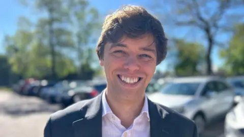 Head and shoulders image of Cameron Holloway. He has brown hair and is wearing a dark suit and light shirt. He is standing in a car park with a number of vehicles behind him. 