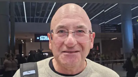Bald man with frameless glasses and light coloured jumper smiles at the camera. In the background people can be seen gathered in an airport arrivals terminal.
