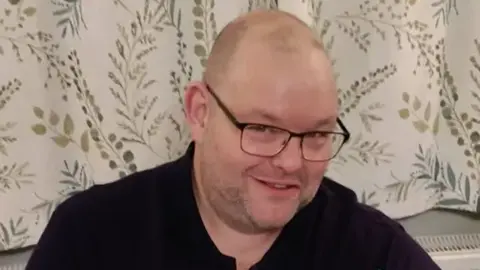 A bald man smiles into the camera. He is wearing black glasses and a black top. Behind him are white curtains with details of green leaves and branches.