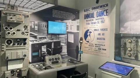 Equipment including a radio control desk with dials and knobs are displayed in front of a wall which includes photos of what the transmission station looked like and a poster for an Annual Dance being held in Droitwich in 1957.