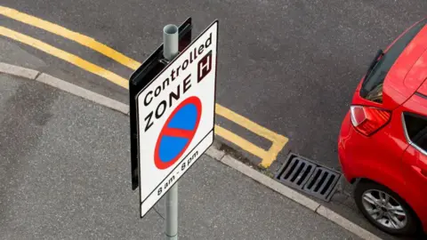 Getty Images A road sign saying "Controlled zone, 8am-8pm" with a red car stopped next to it.