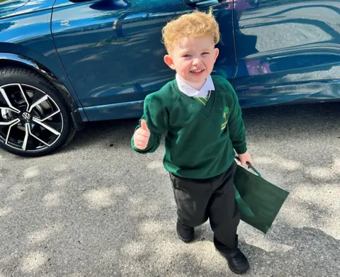 A young boy in a green school jumper with his thumb up