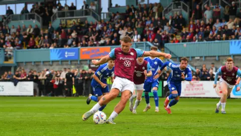 South Shields FC/Kev Wilson Paul Blackett strikes his penalty as fans in the stands look on. Players from both sides are beginning to run towards goal behind him.