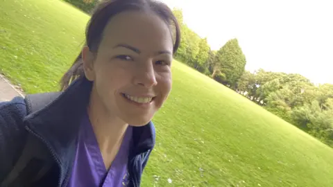 Naomi Durnham A selfie of a woman stood in a park. She has tied-back dark hair and is wearing purple nurse scrubs and a navy fleece. She is smiling and looking at the camera, with green grass and trees behind her.