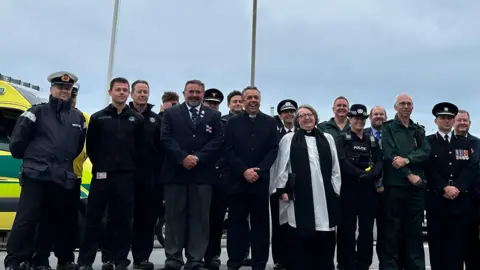 BBC Men and women in different uniforms, including police, fire, paramedics and ecclesiastical, stand with hands clasped in front of two flag poles.