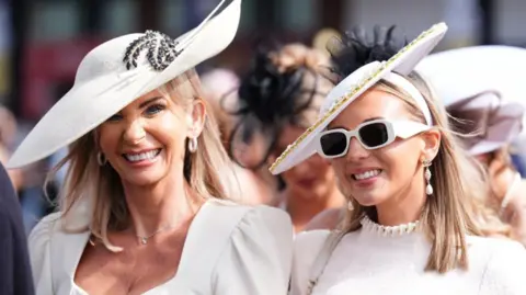Two women wearing white dresses and hats arrive at Aintree