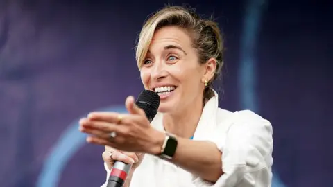 PA Media Close up of Vicky McClure standing on stage with a microphone in her right hand and her left hand stretched out towards the audience, smiling