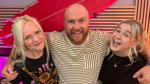 Edward Rowe has his arms around the shoulders of Sarah Buck and Danni Diston. They are all smiling as they stand in the podcast studio. He is wearing a brown and white striped T-shirt and has a bald head and ginger beard. Sarah has long blonde hair and is wearing sunflower patterned dungarees over a black T-shirt. Danni also has long blonde hair and is wearing a black top with a gold necklace. 