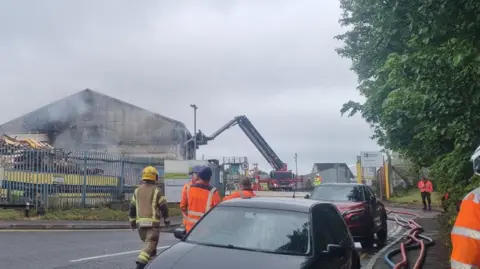 DWFRS A fire engine with aerial ladder next to a smoking building. Hose cables line the pavement.