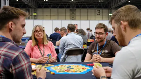 UK Games Expo Three men with brown hair and a woman with blonde hair sitting around a table with a blue hexagonal board between them and a number of cards and counters laid out around it