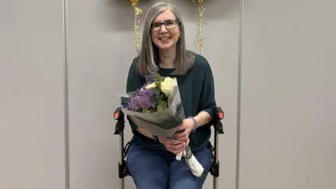 Jo Cole A person sits on a chair indoors, holding a bouquet of flowers wrapped in paper. The bouquet contains white roses and purple flowers, with green foliage. Gold decorations hang in the background against a plain light-coloured wall.