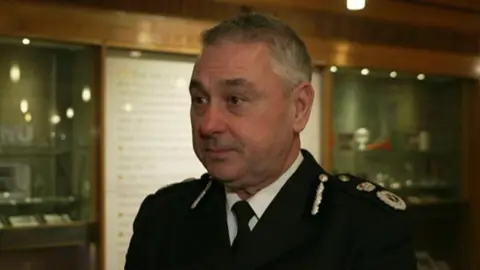 James Vaughan looking to the left. He is stood inside of a building and is wearing a black blazer with badges on the right shoulder.