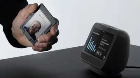 A man's hand shown holding a prototype of Microsoft's new "wearable access badge" -- a small black square with a touch screen about the size of an ID card -- next to a small desktop cube with an interactive screen. 