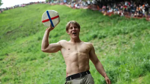Reuters Tom Kopke stands at the foot of Cooper's Hill holding the wheel of cheese aloft. He is bare-chested and wears khaki trousers. There are deep grazes on his sides.