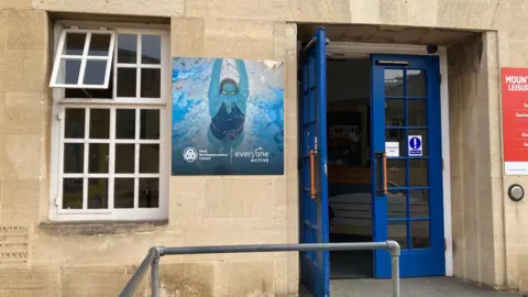Alex Pope/BBC The outside of a stone built swimming pools, with blue open doors, and a sign to the left of the doors and a window to the right. A metal railing is also outside the building.