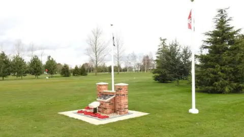 Adrian S Pye/Geograph A memorial bearing the details of the airmen who died, with a British and Canadian flag flying at either side, on a green lawn on a golf course. There are poppy wreaths on the ground in front of the memorial.