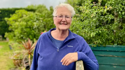 A woman in a purple jumper is standing in a garden area. She is leaning over a wooden fence. Trees and bushes are in the background. 